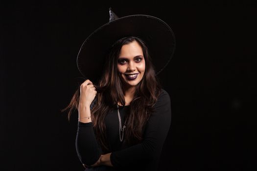 Young beautiful witch with a black dress and a big hat smiling over black background. Pretty girl ready for halloween dressed up like a witch.