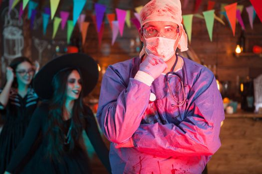 Portrait of suprised man dressed up like a doctor for halloween celebration. Scary witch in the background.