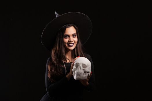 Malefic witch woman with a big hat looking into the camera with a human skull in her hands. Pretty young girl dressed up like a witch for halloween