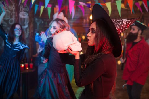 Attractive woman dressed up in a witch costume kissing a human skull at halloween gathering. Scary witch.