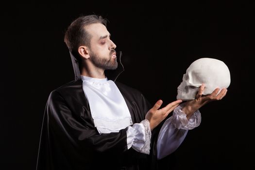Man dressed in Dracula costume for halloween looking at a human skull. Man with vampire teeth. Spooky man with halloween costume.