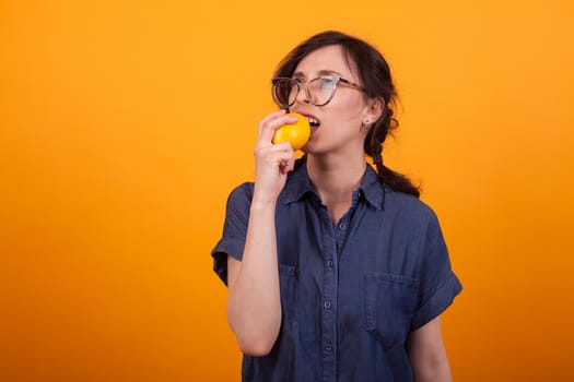 Portrait of beaufitul confused young woman while bitting a fresh orange over yellow background. Girl with juicy orange.
