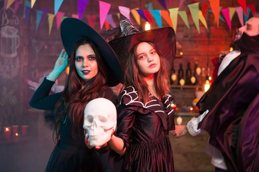 Women in witch halloween costumes standing with a human skull in their hand. Celebrating halloween.