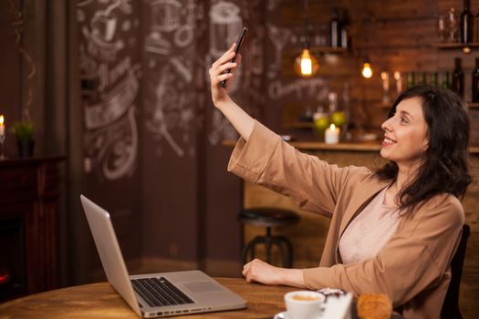 Pretty young girl talking a selfie with her mobile phone in a modern coffee shop. Young female relaxing in a cafeteria during break work. Woman with laptop in front of her.