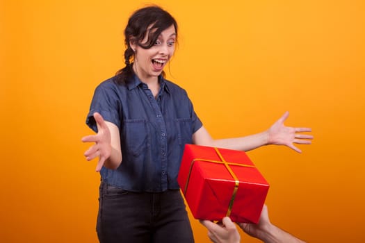 Portrait of cheerful young woman when seeing a red gift box in studio over yellow background. Attractive girl with emotions.