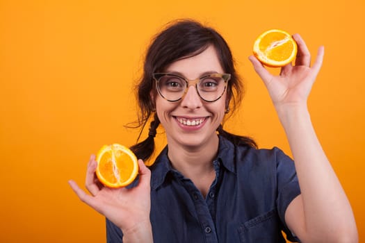 Portrait of attractive young woman with glasses smiling and showing fresh oranges at the camera in studio over yellow background. Cheerful young woman with summer citrus orange. Pretty girl holding jucy oranges .