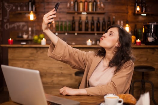 Portrait of attractive young caucasian girl taking selfie in a casual coffee shop. Girl with modern notebook near her in a autehnic coffee shop.