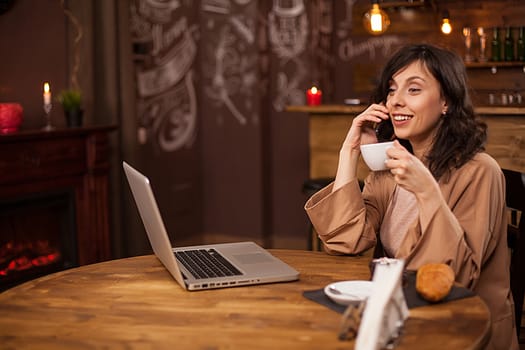 Portrait of caucasian young business woman with compur in a coffee shop. Young female holding coffee cup. Young freelancer talking on smartphone