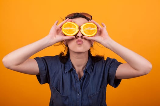 Portrait of funny girl with sliced oranges on her eyes and giving kisses for the camera in studio over yellow background. Cheerful caucasian young woman with fresh oranges. Girl with summer fruits.
