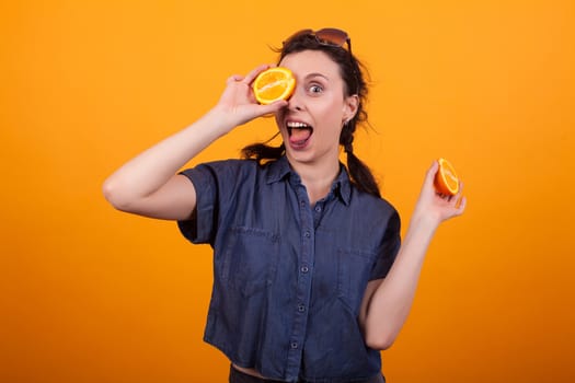 Portrait of a lovely young smiling and covering one of her eye with a fresh and tasty orange in studio over yellow background. Cute lady with delicious citrus. Beautiful woman holding sliced summer fruit.