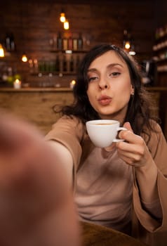 Cheerful beautiful woman giving kissing and taking a self-portrait with smartphone in a coffee shop. Girl holding a cup of coffee. Modern cafeteria.