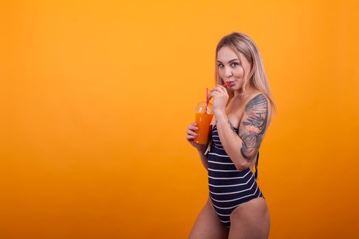 Side view of confident blonde in blue swimming suit and tattoo on arm and drinking refreshing cocktail over yellow background. Young woman with sexy body,