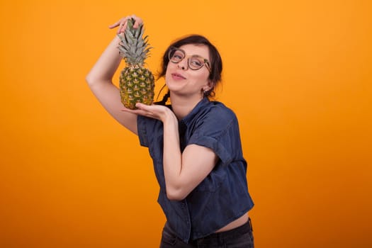 Excited young beautiful woman with glasses happy about her delicious pineapple in studio over yellow background. Beautiful woman with exotic fruit. Pretty girl holding a pineapple.