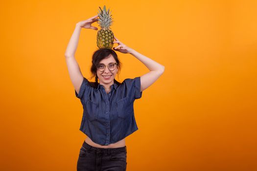 Young sexy woman happy smiling with glasses holding a tasty pineapple on her head in studio over yellow background. Cheerful woman holding delicious ananas.