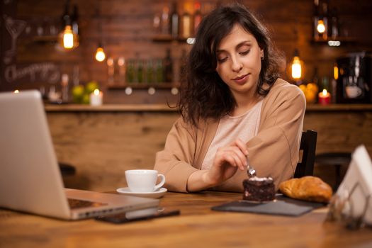 Beautiful woman eating delicious chocolate cake in a coffee shop. Girl in front of laptop in a coffee shop. Girl enjoying tasty cake.