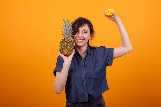 Laughing young beautiful woman with tropical fruits in studio over yellow background. Tasty ananas. Delicious pineapple. Summer fruits.