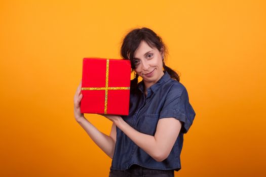 Portrait of happy young woman with her red gift box looking at the camera in studio over yellow background. Cute young woman with gift box. Red gift box.