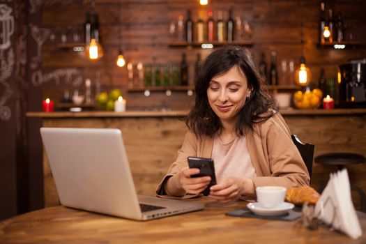 Portrait of a beautiful woman sitting in a coffee shop texting on her phone. Laptop witt wireless internet connection. Cup of coffee. Smart young woman. Woman writing a message on her phone. Beautiful girl smiling.