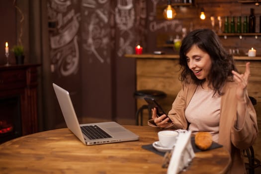 Portrait of modern female entrepreneur using smartphone in a conference call. Woman with modern laptop near her in coffee shop. Coffee shop with wireless internet connection. Smiling beautiful woman in a coffee shop.