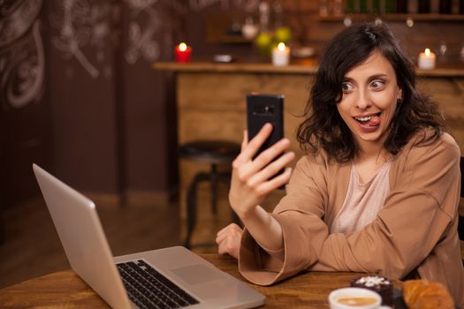 Smiling beautiful young woman taking funny selfie on her phone sticking her tongue out in a coffee shop. Girl with lapto near her. Funny lifestyle.