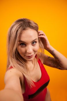 Beautiful young woman with fit body in swimming suit taking a selfie and playing with her hair over yellow background. Charming young woman in swimming suit. Sexy young woman.
