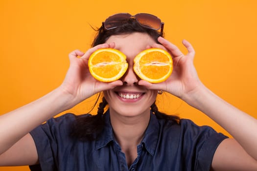 Portrait of cheerful young woman with sliced orange as eyes in studio over yellow background. Pretty caucasian young woman holding a sliced orange.