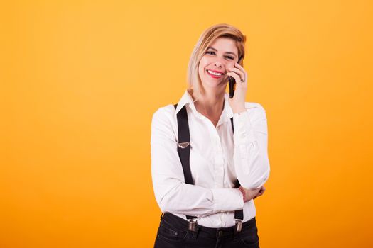 Pretty young woman looking at the camera while talking on the phone over yellow background. Hipster clothes. Attractive woman.
