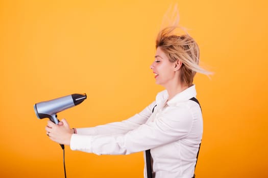 Young funny woman blowing her short hair with a hair drayer over yellow background. Cool hairstyle. Haircare.