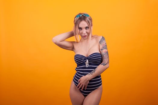 Sexy young tattooed woman in blue swimming suit over yellow background. Charming woman in swimming suit and swimming glasses ready for vacation on the beach .