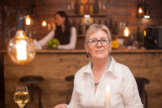 Portrait of elderly woman in a vintage restaurant. A glass of white wine. Blurred barmaid working in the background.