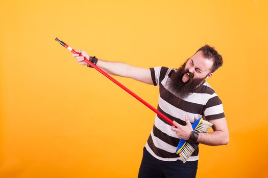 Funny bearded rockstar playing virtual guitar with broom over yellow backgrounnd. Handsome man. Cheerul man.