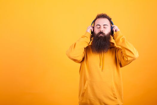 Bearded young man wearing yellow hoodie ,listening music on his blue headphones peacefully over yellow background. Handsome man. Great song.