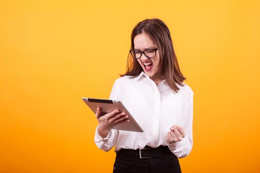 Pretty young teenager can't contain her happines and holding a tablet in studio over yellow backgroud. Caucasian girl using tablet. Girl using modern technology.