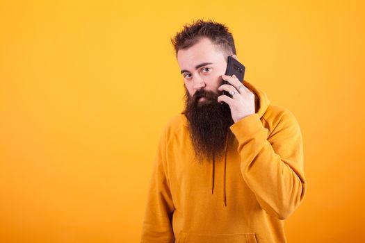 Bearded guy looking worried at the camera while talking on the phone over yellow background.