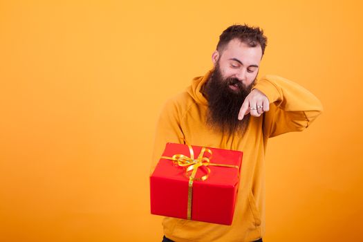 Handsome bearded man in yellow hoodie pointing on red gift box over yellow background. Handsome man. Surprise gift.