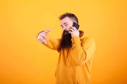 Trendy man wearing yellow hoddie talking on the phone and showing ok sign over yellow background. Smartphone. Stylish man.