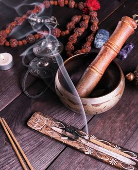 copper singing bowl and a wooden stick on a brown table, a stick with incense is burning nearby