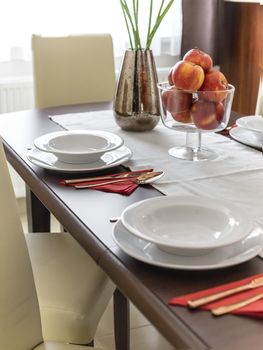 Tableware and a big bowl of fruit on a dining table in natural light, angled view.