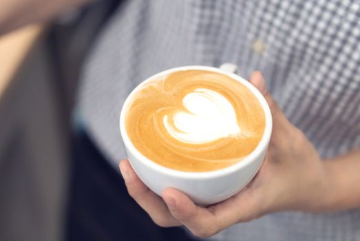 Woman barista holding latte art coffee with heart shape in white cup, selective and soft focus