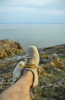 Feet of male tourist resting on the rocks at the beach at sunset, looking at the water.