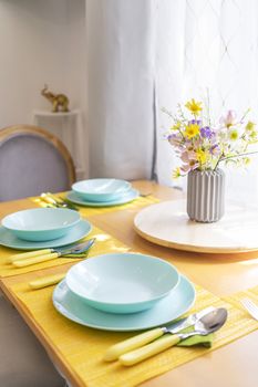 Colorful set of tableware and a bouquet of wild flowers on a brown wooden dining table in natural light, angled view.