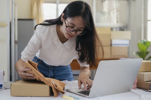 Young entrepreneur working online e-commerce shopping at her shop. Young online seller checking preduct for prepare parcel box deliver to customer. Online selling and online business e-commerce concept.