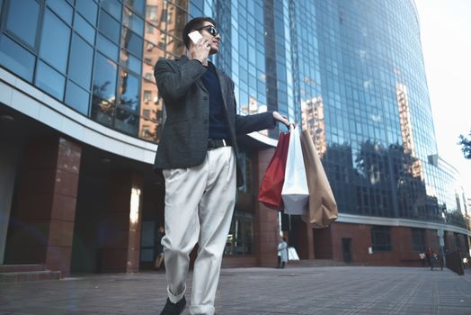 Positive cool guy with sunglasses and paper bags walking near showcase with reflection.