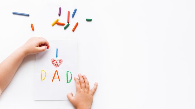 Left-handed toddler draws greeting card for Father's day. Kid uses wax crayons to write I Love Dad and to paint red heart symbol.Top view on child's hands,pencils on white background with copy space.