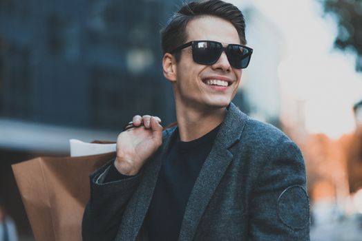 Positive cool guy with sunglasses and paper bags walking near showcase with reflection.