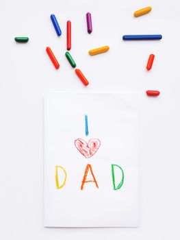 Top view on hand drawn greeting card for Father's day. Kid uses wax crayons to write I Love Dad and to paint red heart symbol. Flat lay with pencils on white background.