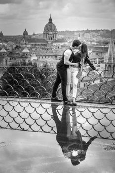 Romantic couple in Rome city, Italy. A beautiful pair embrace on a terrace with a reflection in a puddle. Behind them the historical and ancient city of Rome, Italy. Black and white