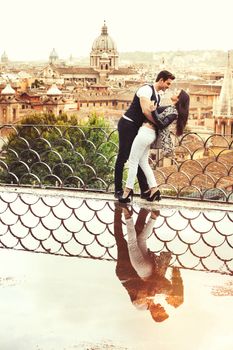 Romantic couple in Rome city, Italy. Loving relationship. Passion and love. A beautiful pair embrace on a terrace with a reflection in a puddle. Behind them the historical ancient city of Rome, Italy.