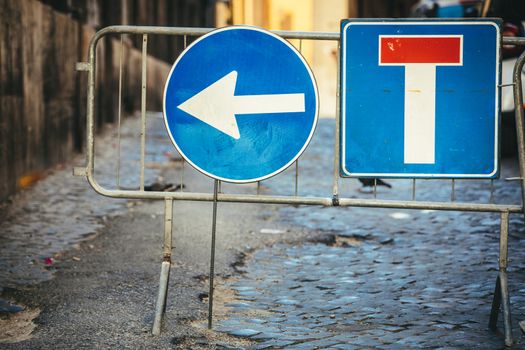 Road works detour road signs. Dead end road and arrow to turn left. Iron crossing barrier. Street with paving cobblestones.