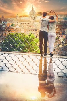 Beautiful pair embrace on a terrace with a reflection in a puddle. Behind them the historical and ancient city of Rome, Italy. Love, passionate affair, youth and genuine love concept.